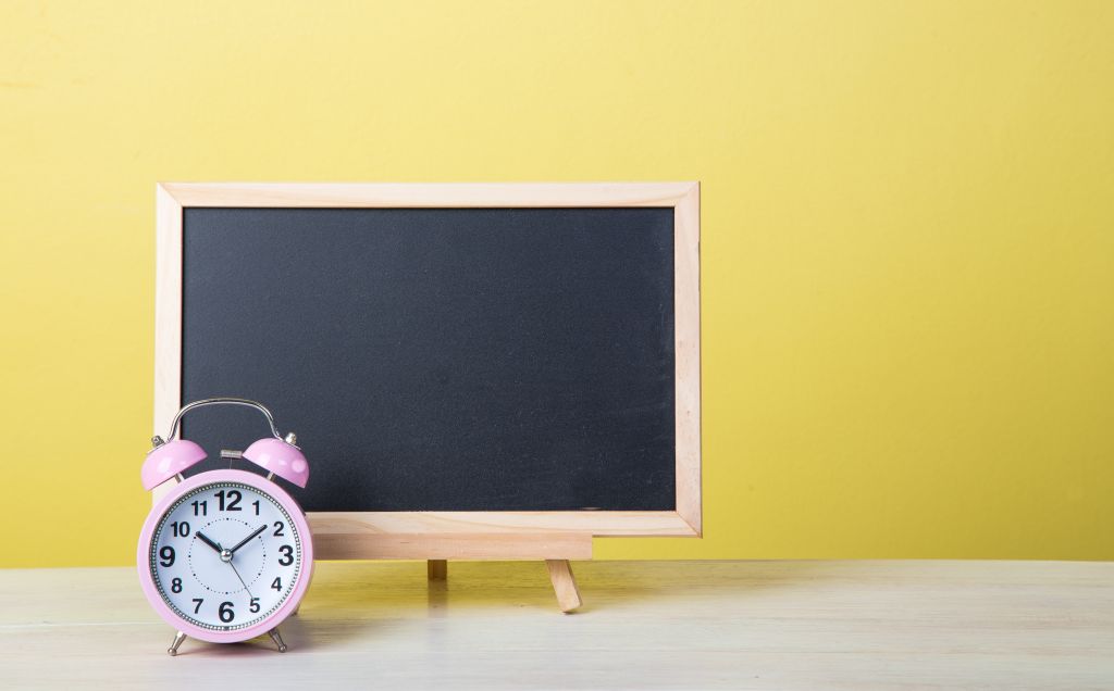 Blackboard And Alarm Clock On Table Against Yellow Wall